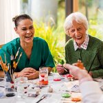 Eine Pflegerin und eine Seniorin sitzen lachend am Maltisch. 