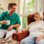 Ein Pfleger unterhält sich mit einer Seniorin bei einer Tasse Kaffee. 