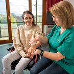 Eine Pflegerin massiert mit einem Igelball das Handgelenk einer Frau im Rollstuhl. 