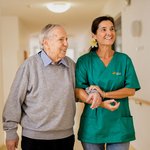 Ein Senior geht lächelnd mit einer Pflegerin durch einen Gang, sie hält seinen Arm.