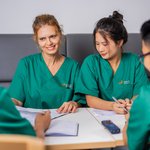 Vier Pfleger*innen in grünen Kasaks sitzen an einem Tisch und besprechen etwas. Zwei sind frontal in der Bildmitte zu sehen, die anderen beiden sind an den Bildrändern von hinten angeschnitten.