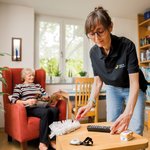 Eine Mitarbeiterin staubt einen Tisch ab, während eine Seniorin im Sessel sitzt und liest.